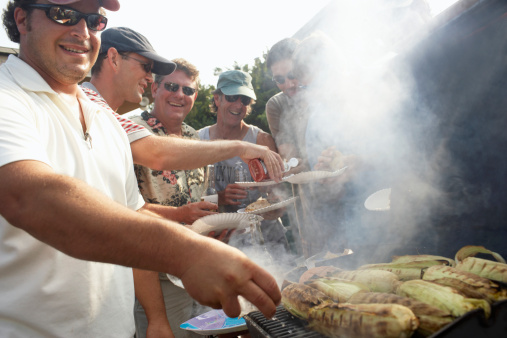Man serving sweetcorn from smoking barbecue, friends in background